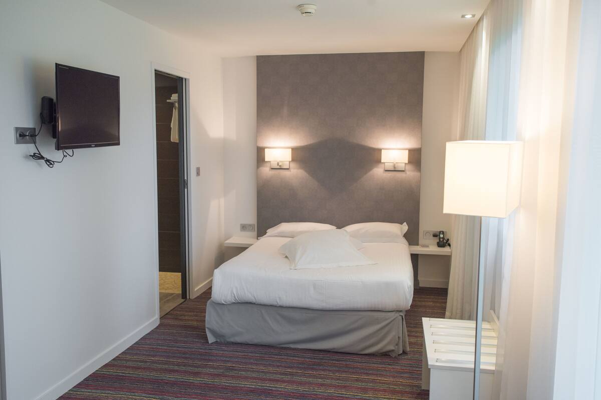 Habitación de hotel con una cama, televisor montado en la pared, dos lámparas de pared, una mesita de noche y una lámpara.