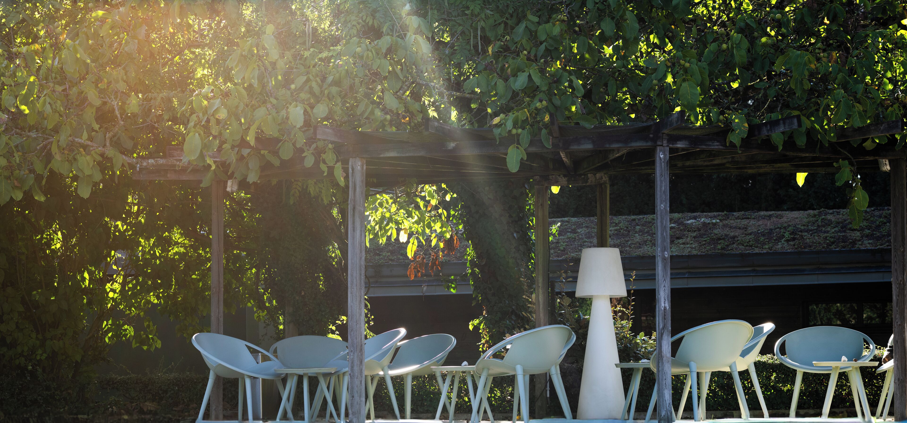 Un espacio al aire libre cubierto con sillas de metal y una mesa, rodeado de vegetación y una estructura de pérgola.