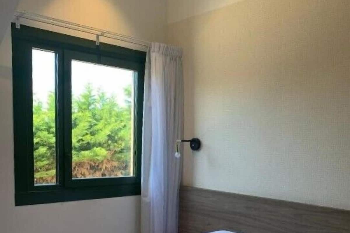Una cama bien tendida con sábanas y almohadas blancas, una ventana con vistas a la vegetación y un interruptor de luz en la pared.