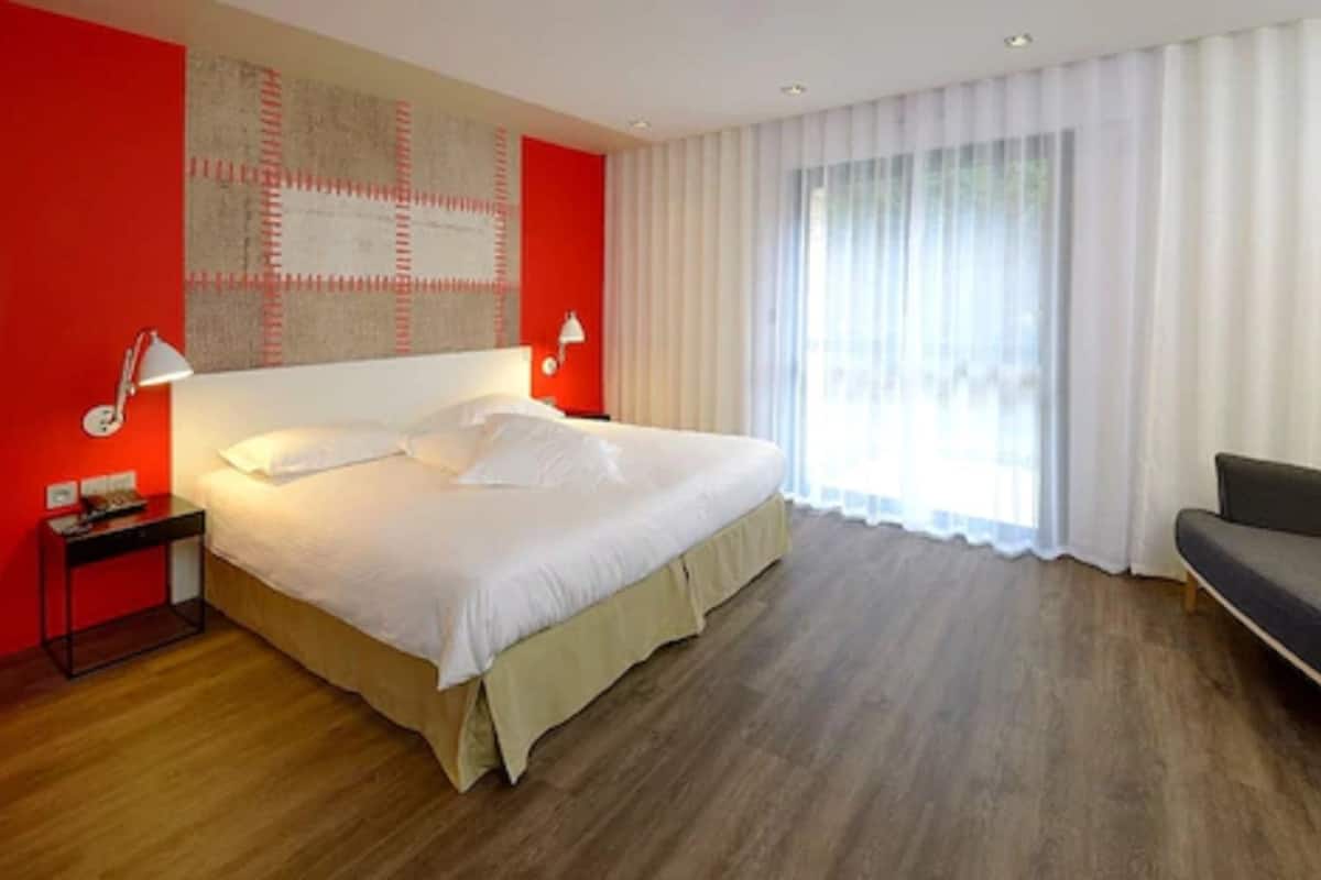 Una habitación de hotel moderna con una cama grande, una mesita de noche, una lámpara de escritorio, un sofá y un amplio ventanal con cortinas traslúcidas.