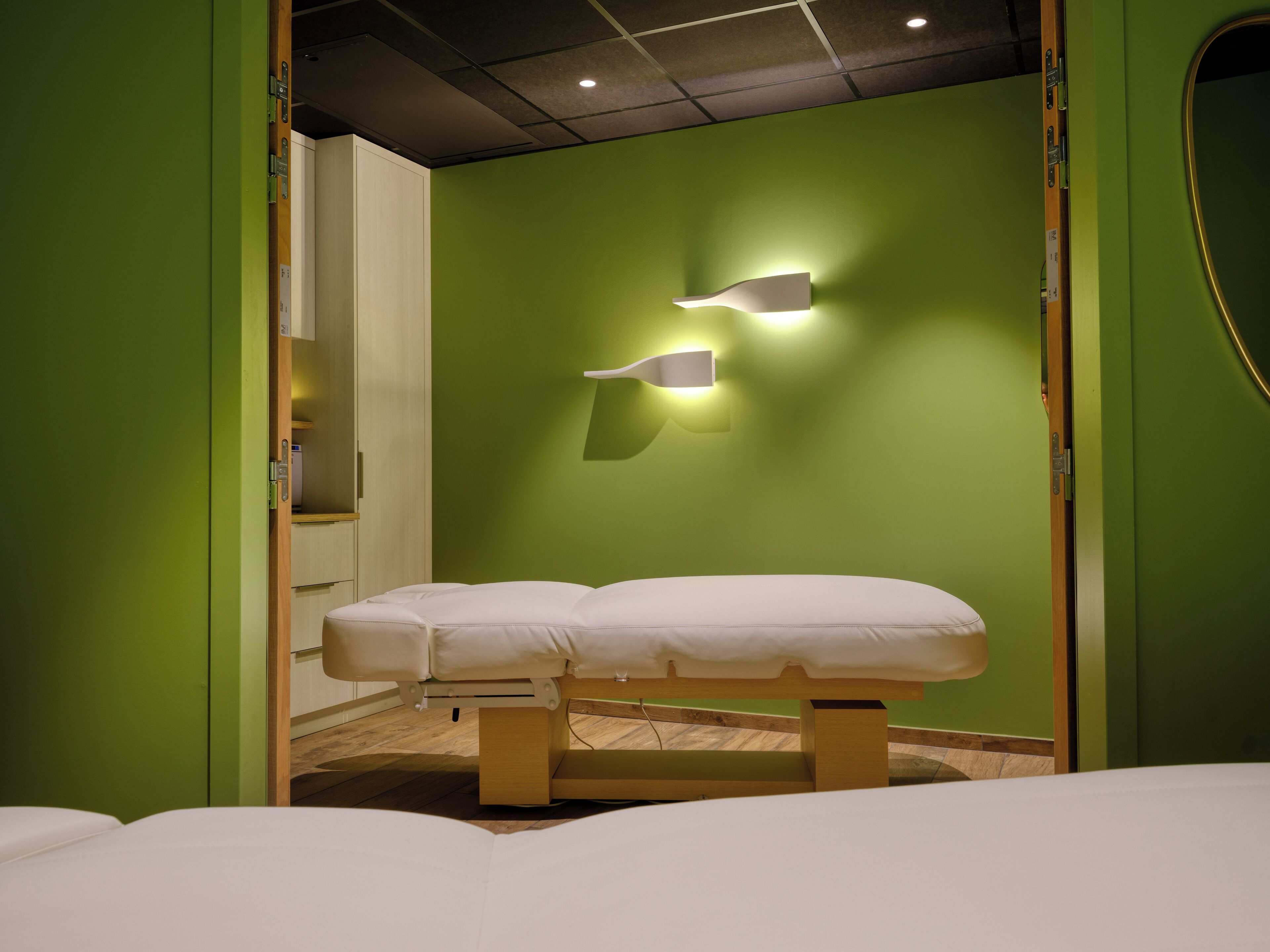 Quarto com parede verde, um banco de madeira com almofada branca e luminária fixa na parede.