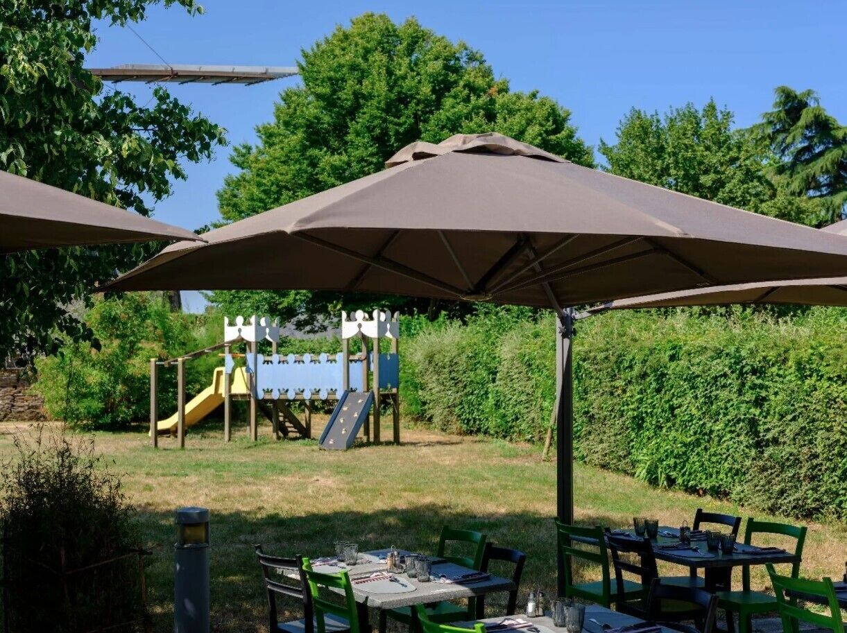 An outdoor patio area with tables and chairs under umbrellas, a children's play structure in the background, and a hedge.