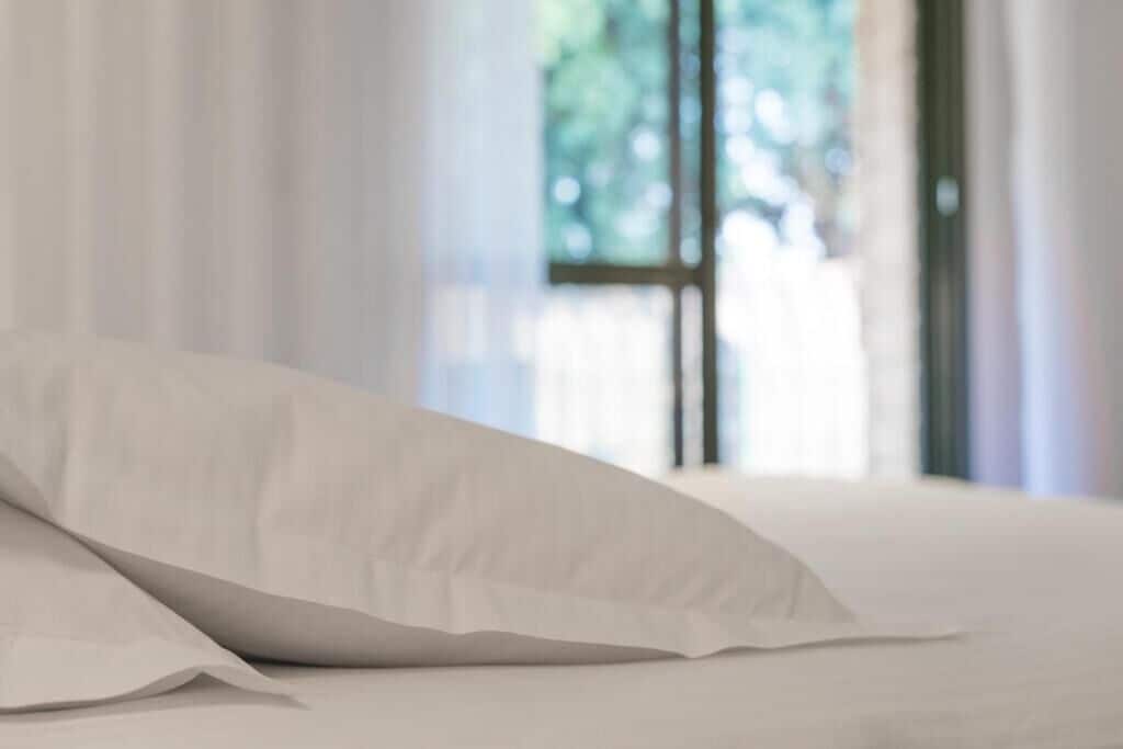 Una cama con ropa blanca y una almohada blanca en una habitación con cortinas translúcidas y una ventana con vistas a la vegetación.
