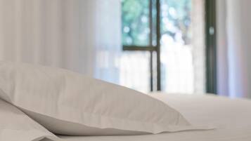 Una cama con ropa blanca y una almohada blanca en una habitación con cortinas translúcidas y una ventana con vistas a la vegetación.