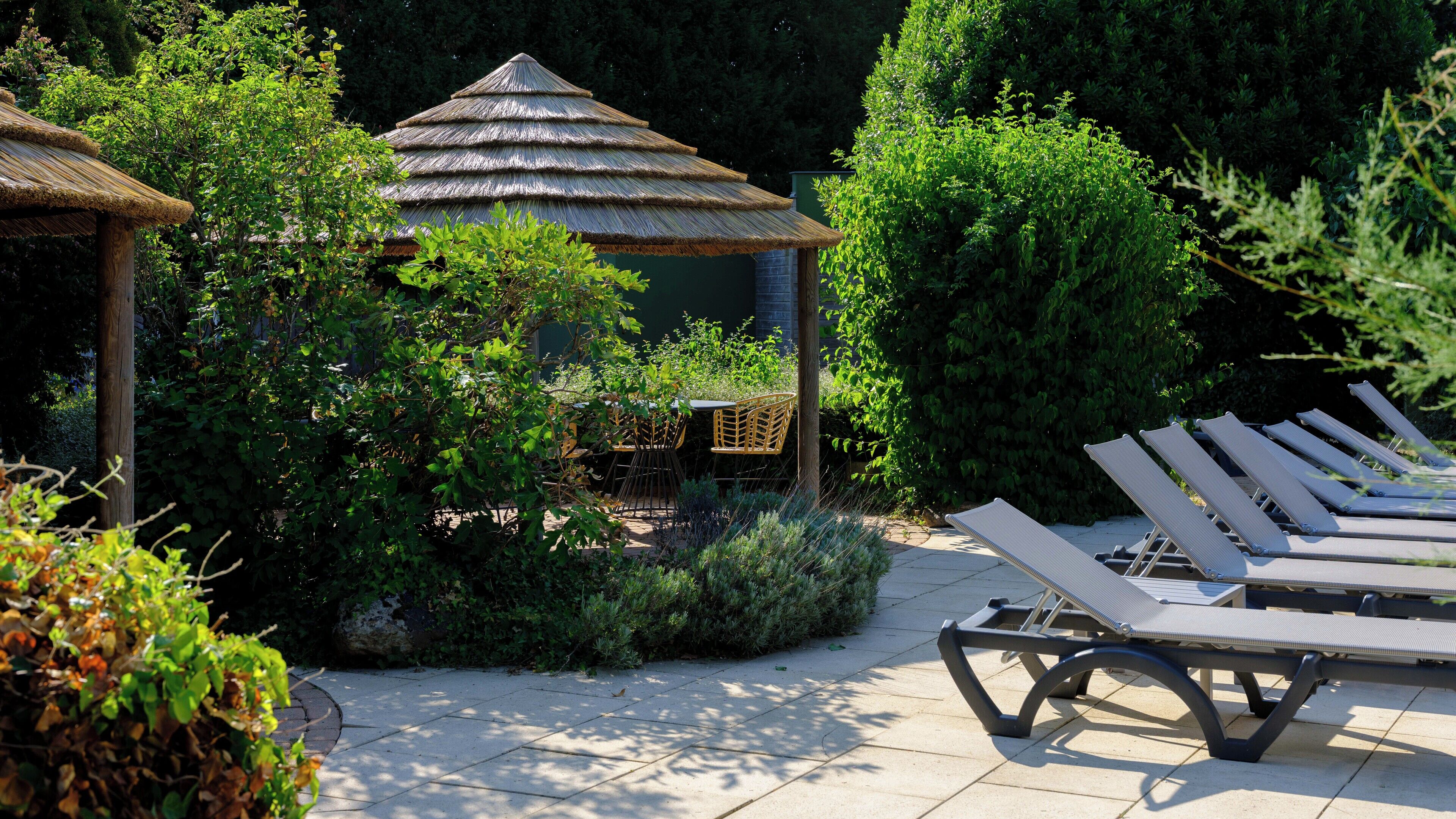 A garden with a thatched gazebo, lounge chairs, and a variety of plants.