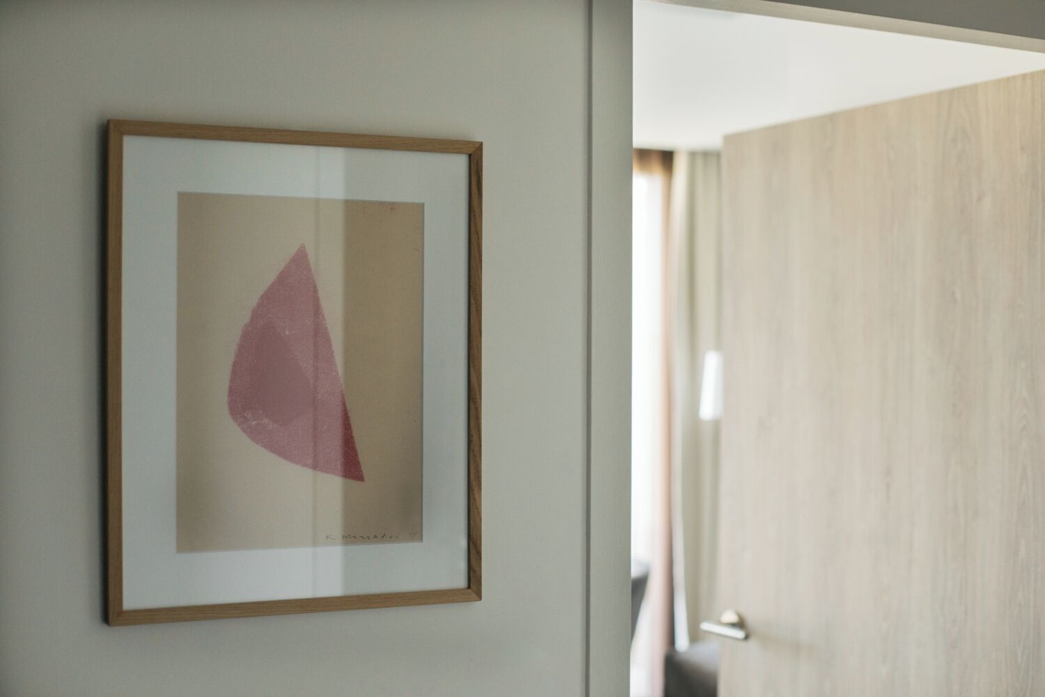 Uma obra de arte abstrata emoldurada numa parede de um quarto com porta de madeira e parede em tom claro.