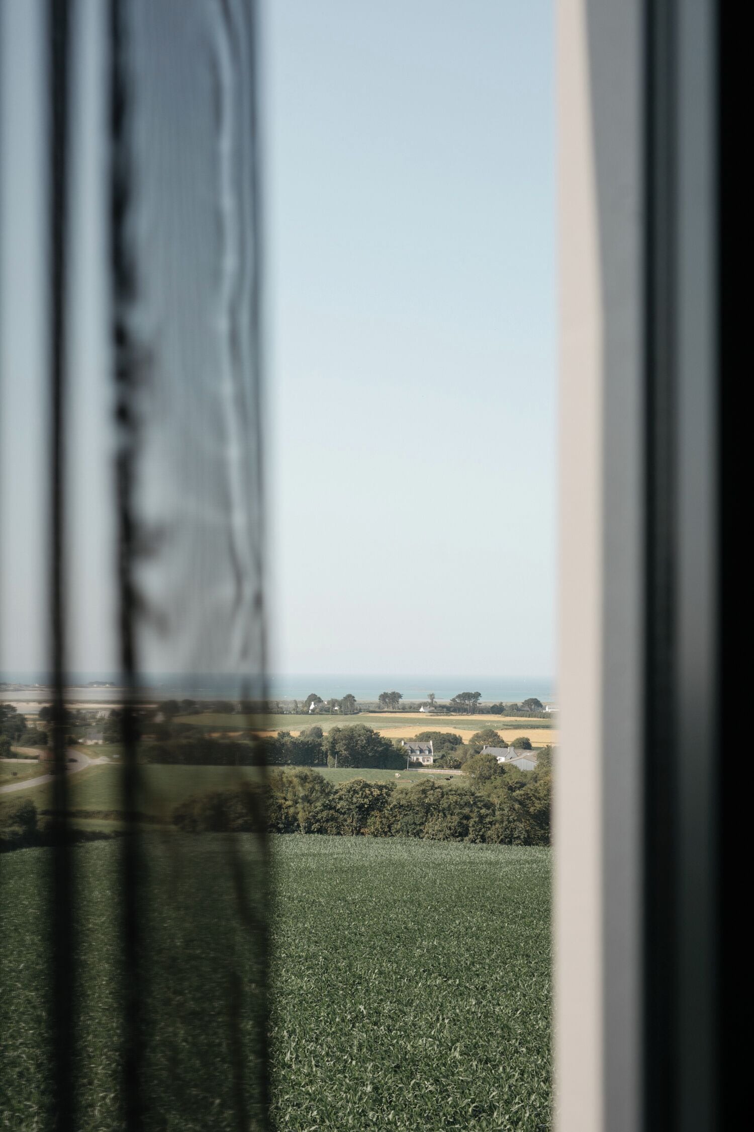 A view through a window with a blurred foreground, revealing a landscape with fields, trees, and distant buildings.