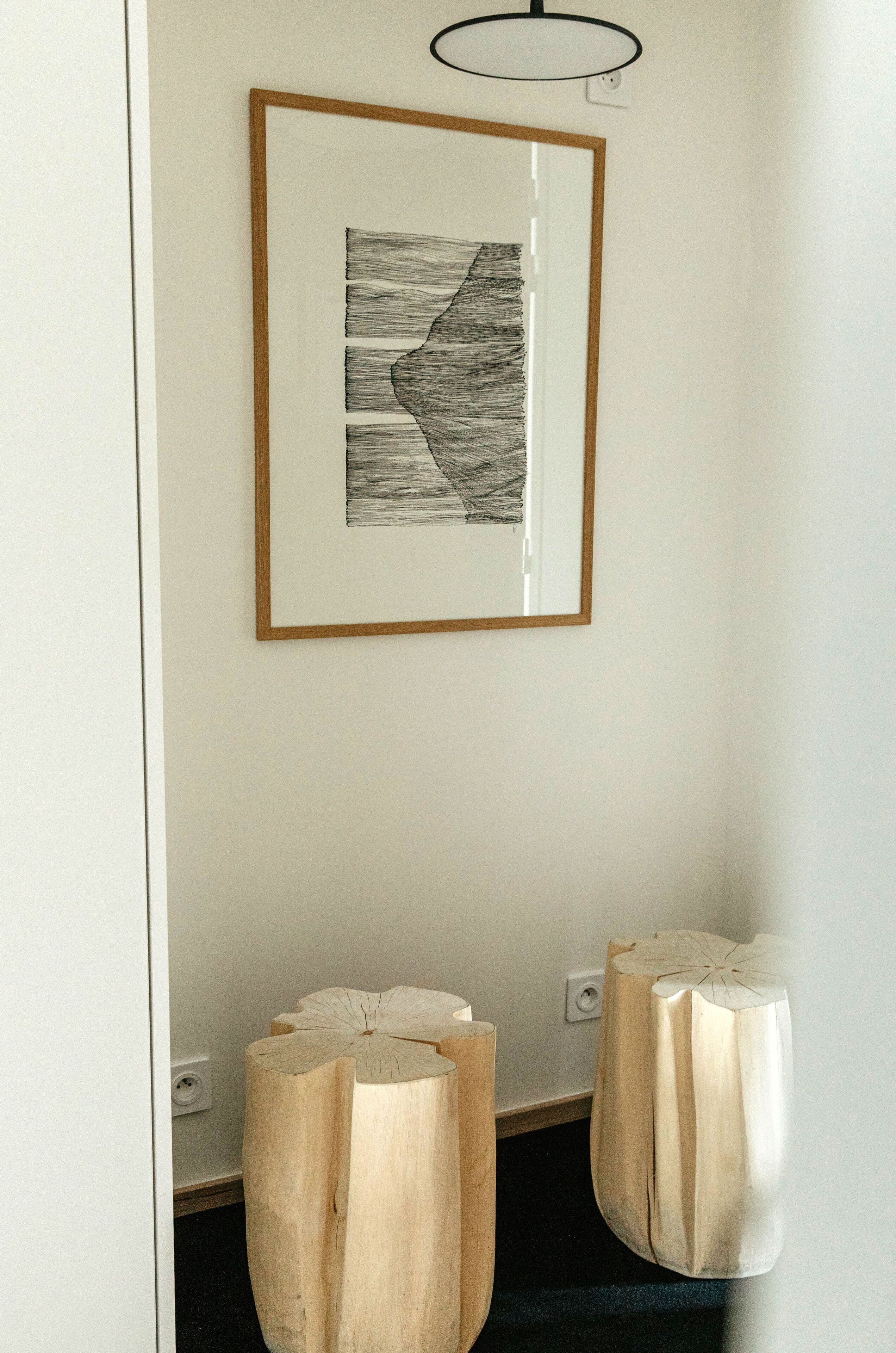 A framed abstract artwork on a wall, wooden stools with a unique design, and a light fixture above the artwork.