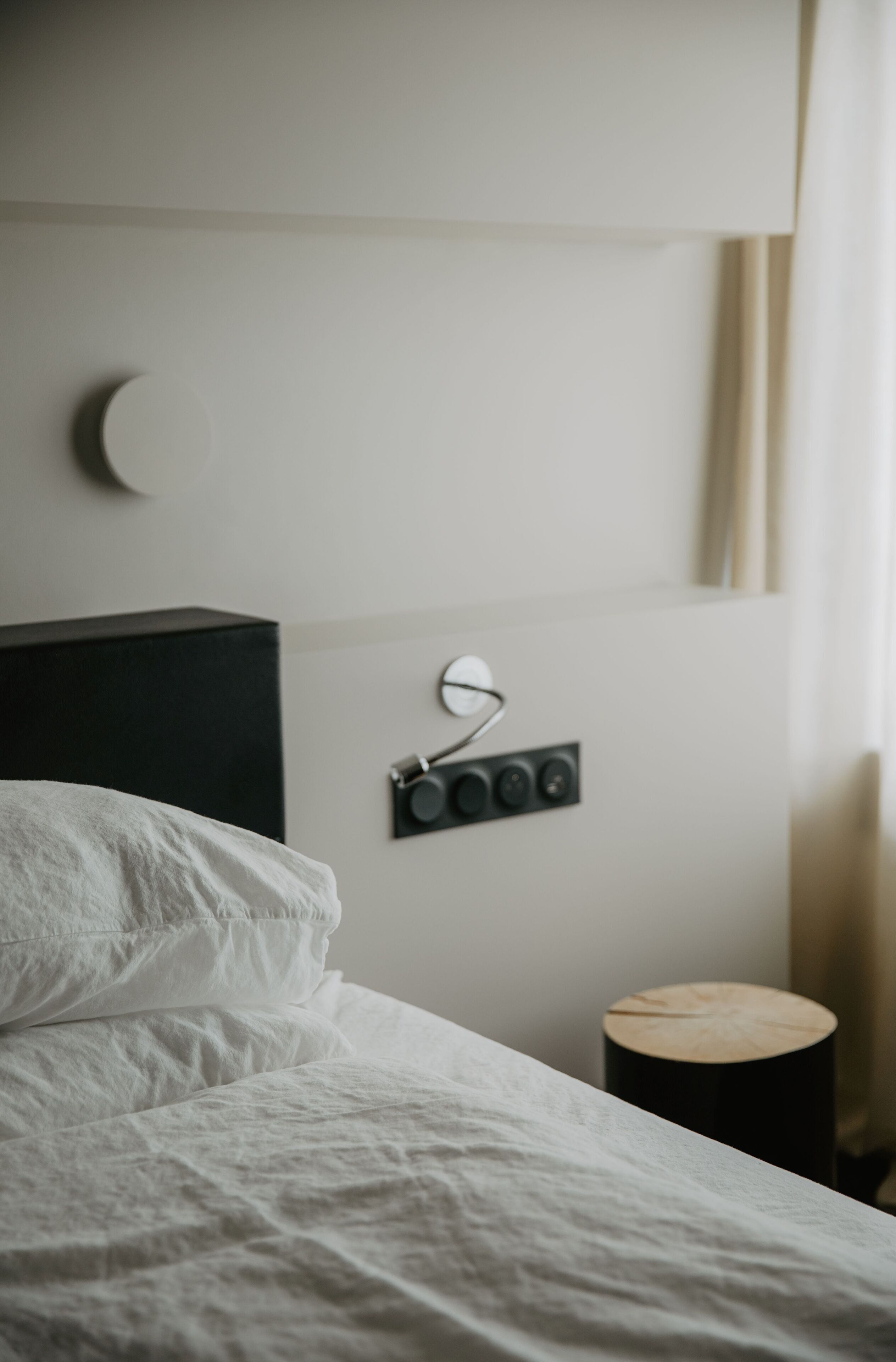 A neatly made bed with white linens, a black headboard, a wall-mounted light fixture, and a small wooden side table.