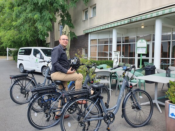 A man sitting on a bicycle in front of a building with a sign for Green Hotels. There are multiple bicycles parked nearby, and a white van is visible in the background. - Green hôtels Parc des Expositions (Tremblay-en-France)