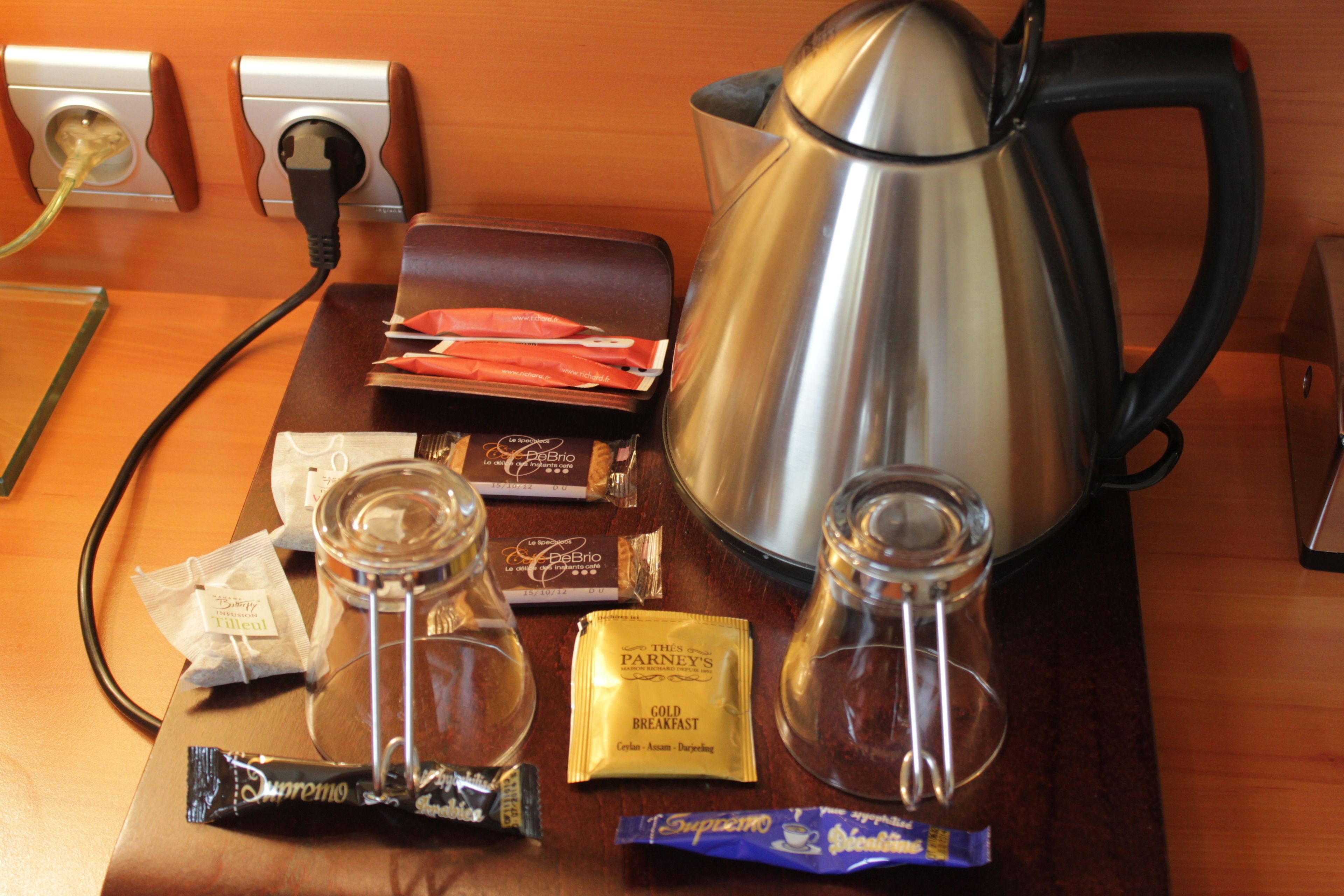Une table en bois, une bouilloire en acier inoxydable, un bocal en verre, une trousse en cuir marron et plusieurs paquets contenant des aliments et des boissons.