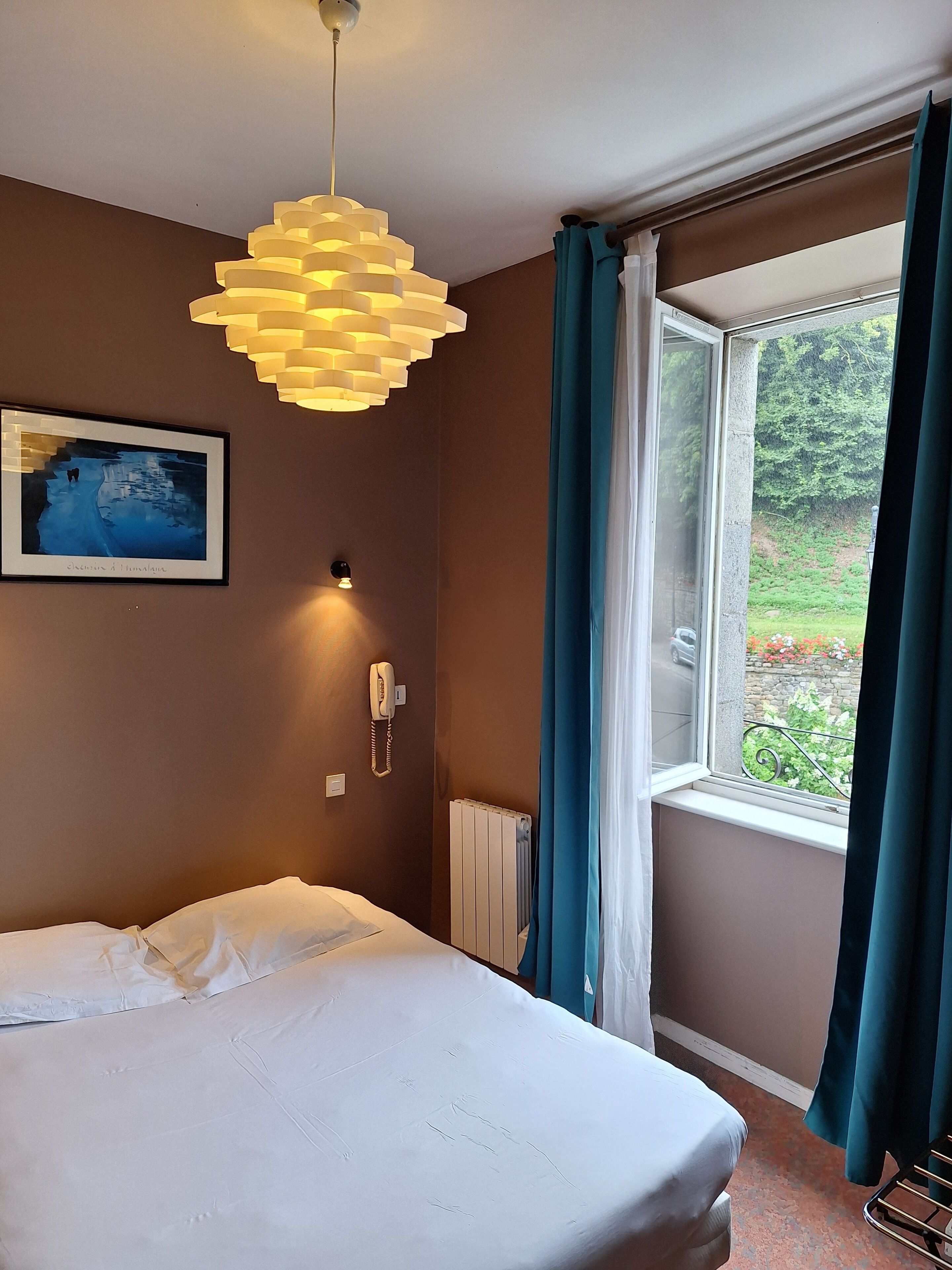 Ein Schlafzimmer mit Bett, einer Pendelleuchte, einem Telefon und einem Fenster mit Vorhängen.
