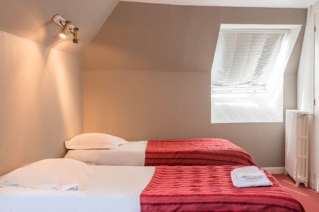 A bedroom with two beds, a window with blinds, a radiator, and a wall-mounted light fixture.