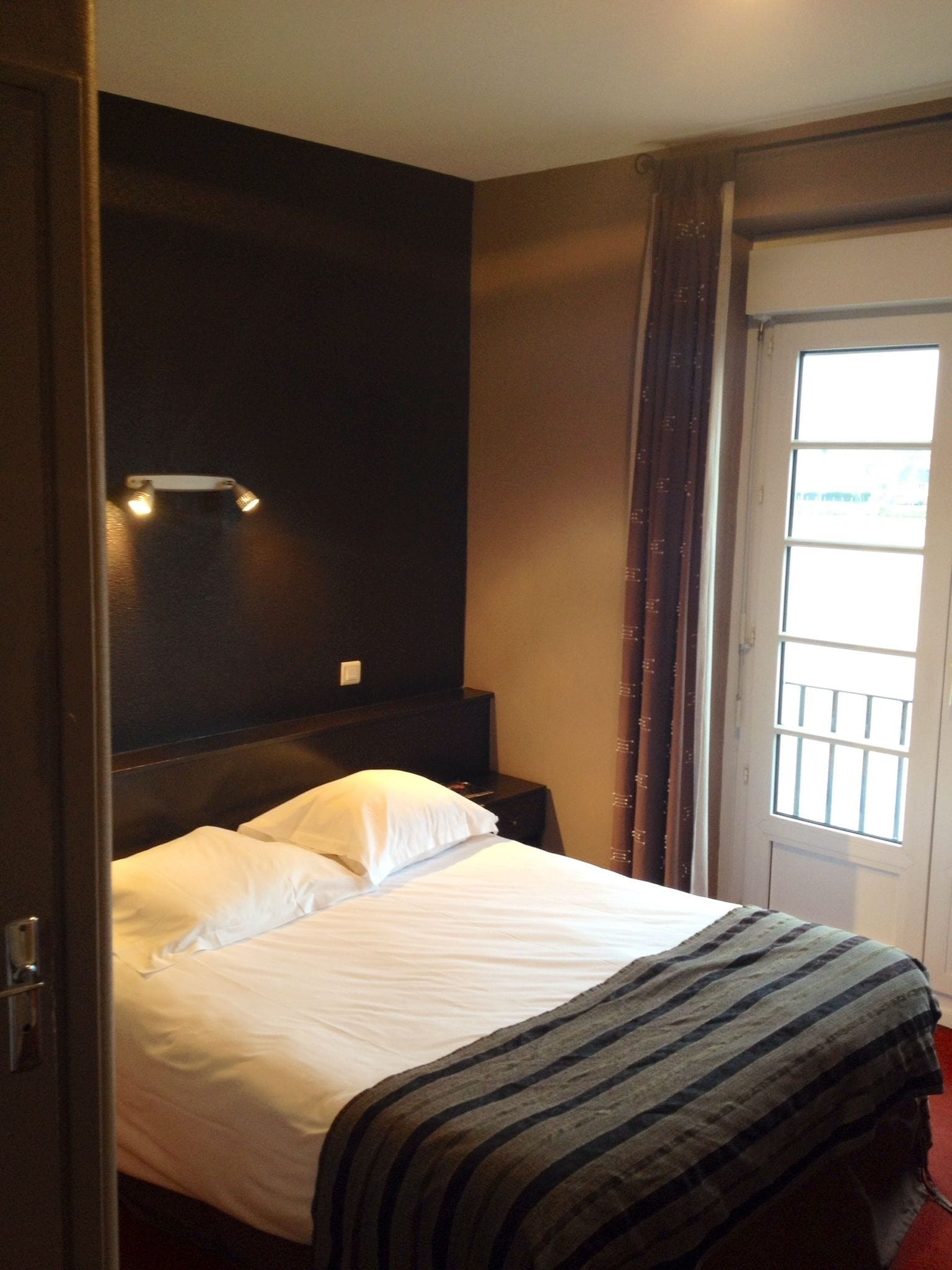A hotel room with a bed, a nightstand, a wall-mounted light, and a window with curtains.