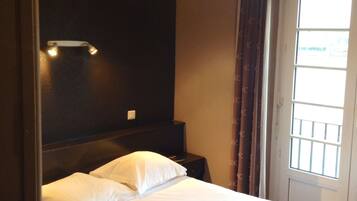 A hotel room with a bed, a nightstand, a wall-mounted light, and a window with curtains.