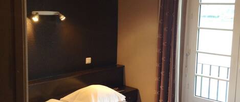 A hotel room with a bed, a nightstand, a wall-mounted light, and a window with curtains.