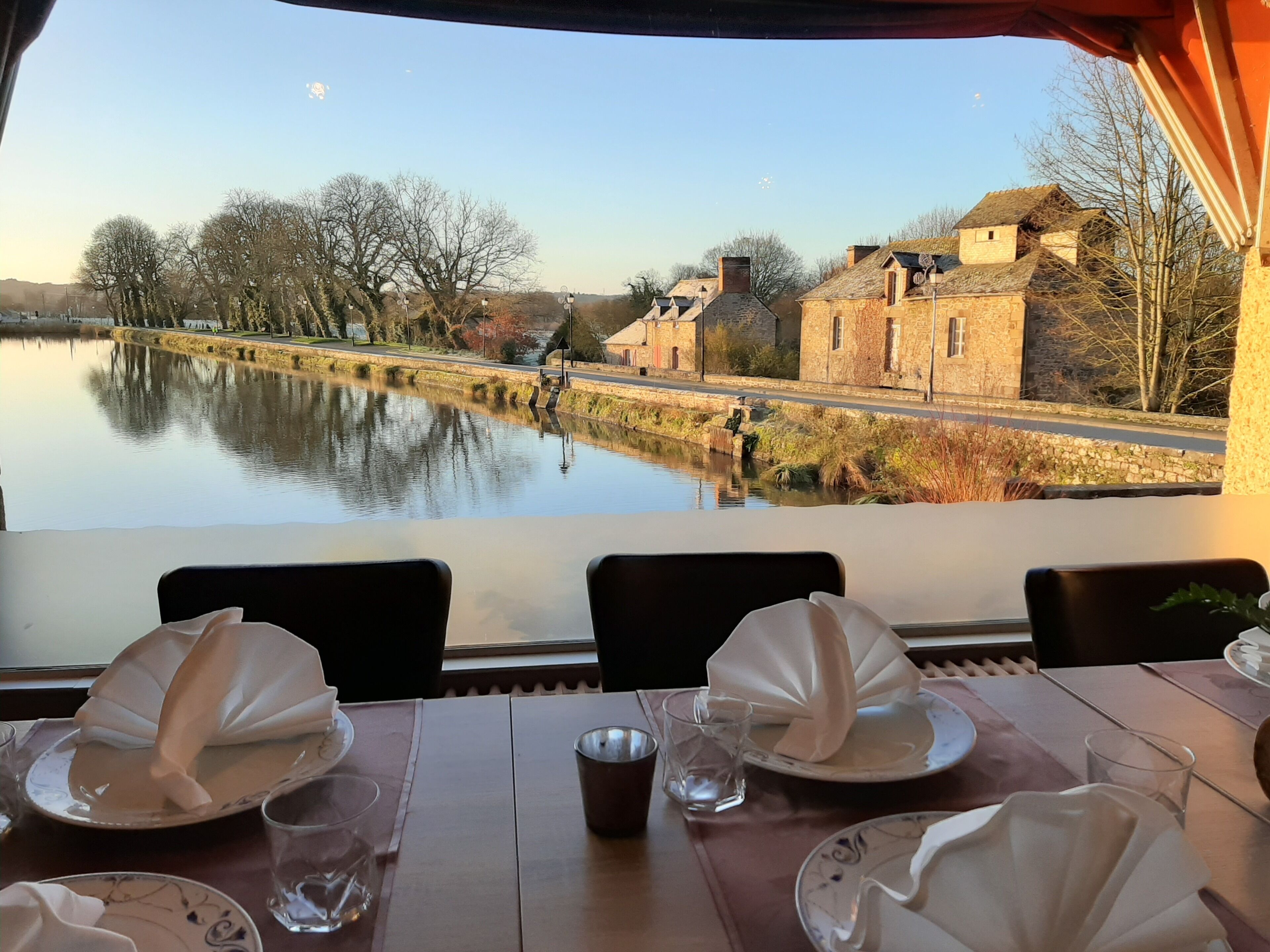 A dining table with plates, glasses, and napkins set for a meal. A window reveals a calm river and buildings in the background.
