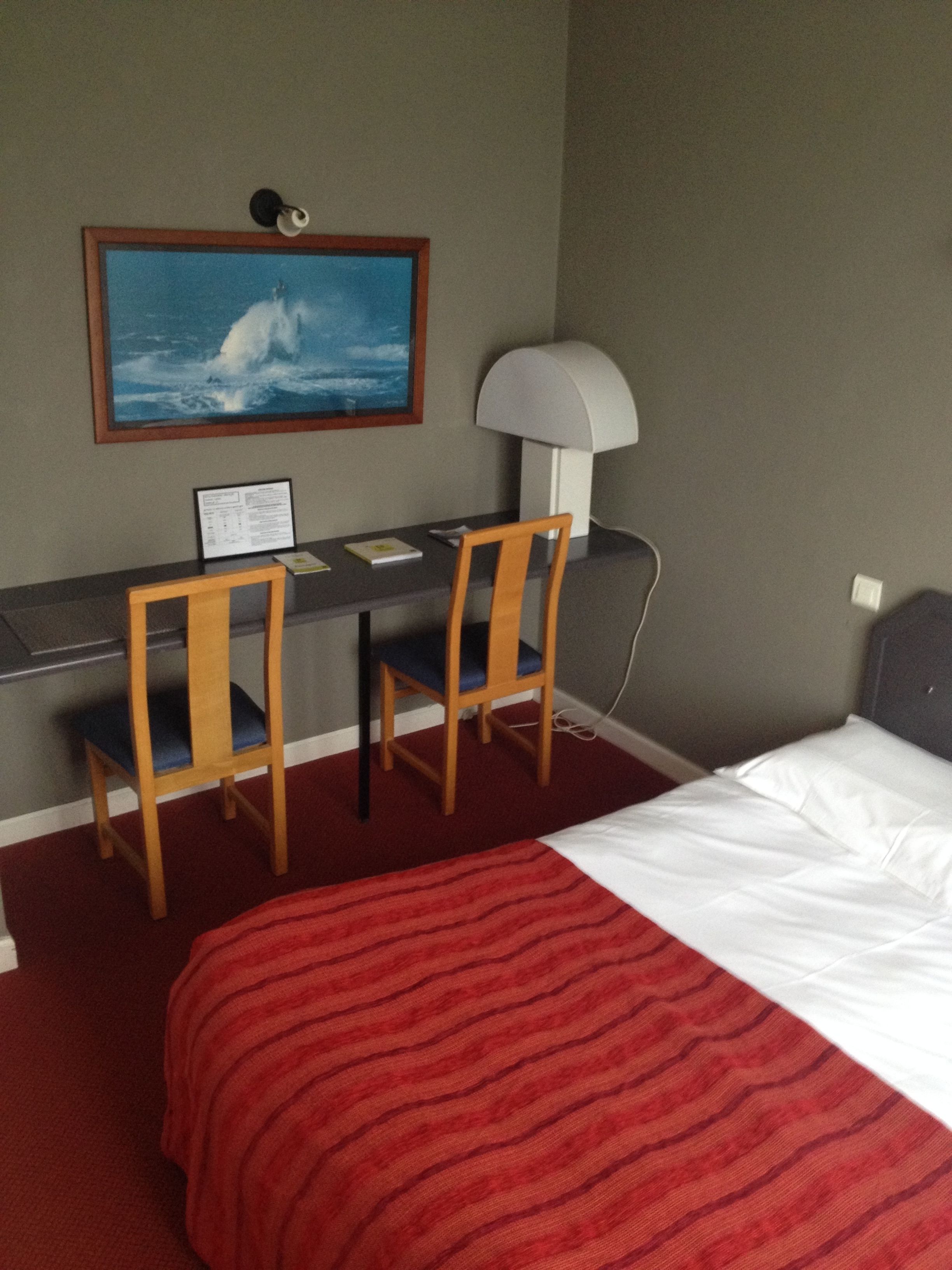 A hotel room with a bed, a desk with two chairs, a lamp, a framed picture on the wall, and a red carpet.