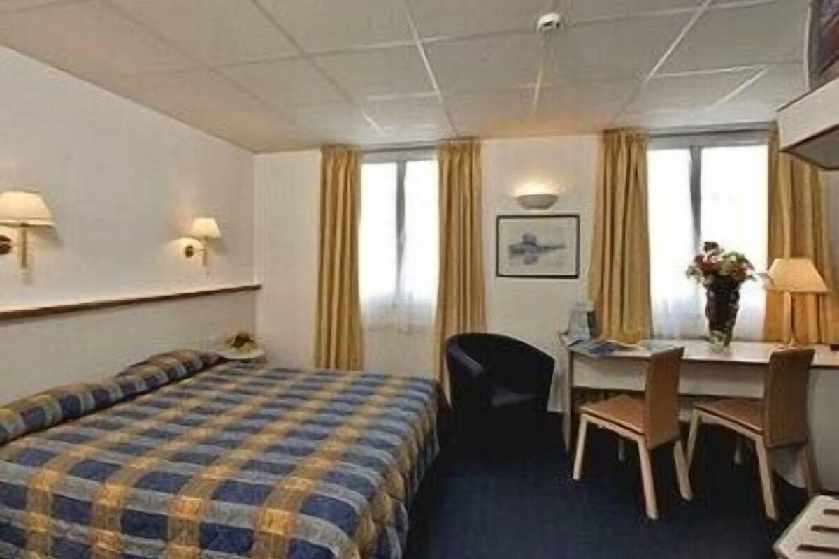 A hotel room with a bed, a desk, a chair, a lamp, and a window with curtains.
