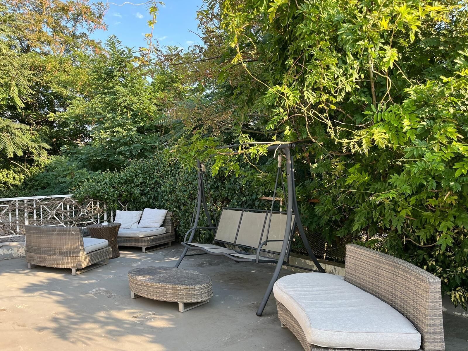 A patio area with wicker furniture, including a sofa, ottoman, and swing set, surrounded by lush greenery.