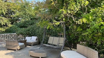 A patio area with wicker furniture, including a sofa, ottoman, and swing set, surrounded by lush greenery.
