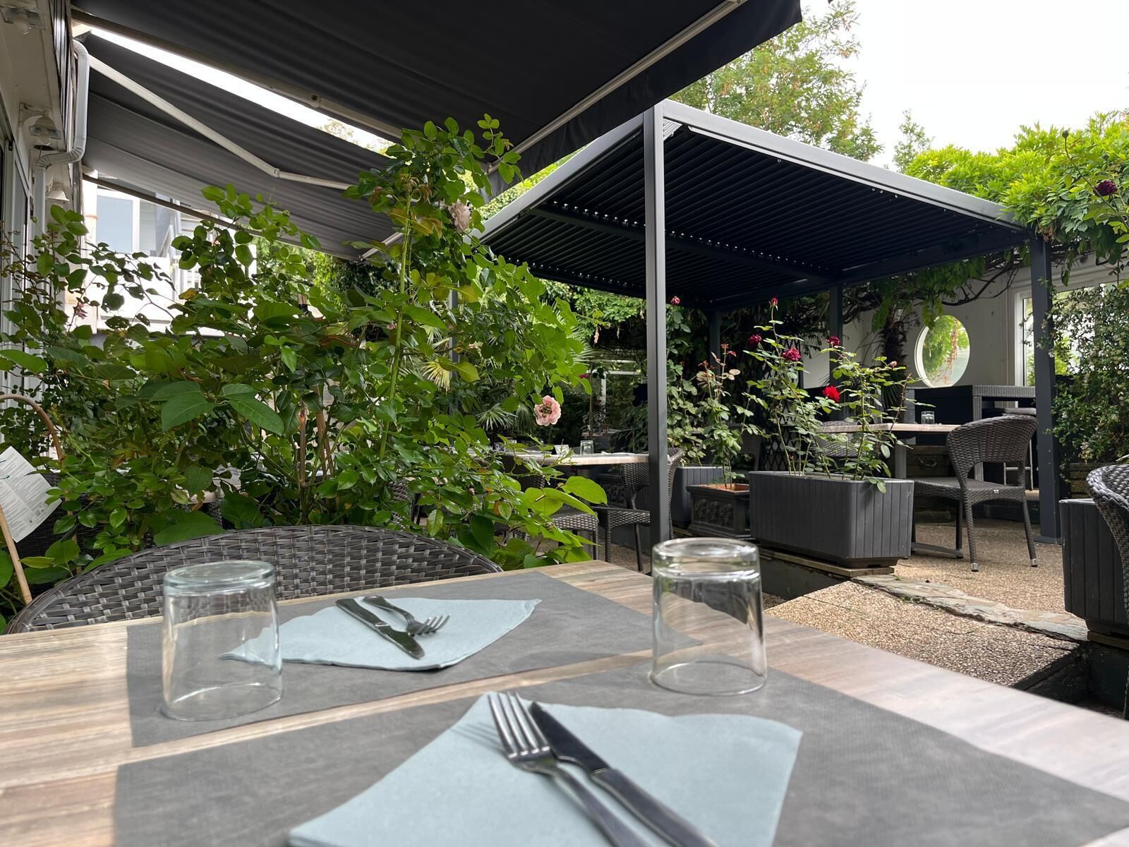 Eine Terrassenfläche im Freien mit Tischen und Stühlen, umgeben von Grünpflanzen und einem überdachten Sitzbereich.