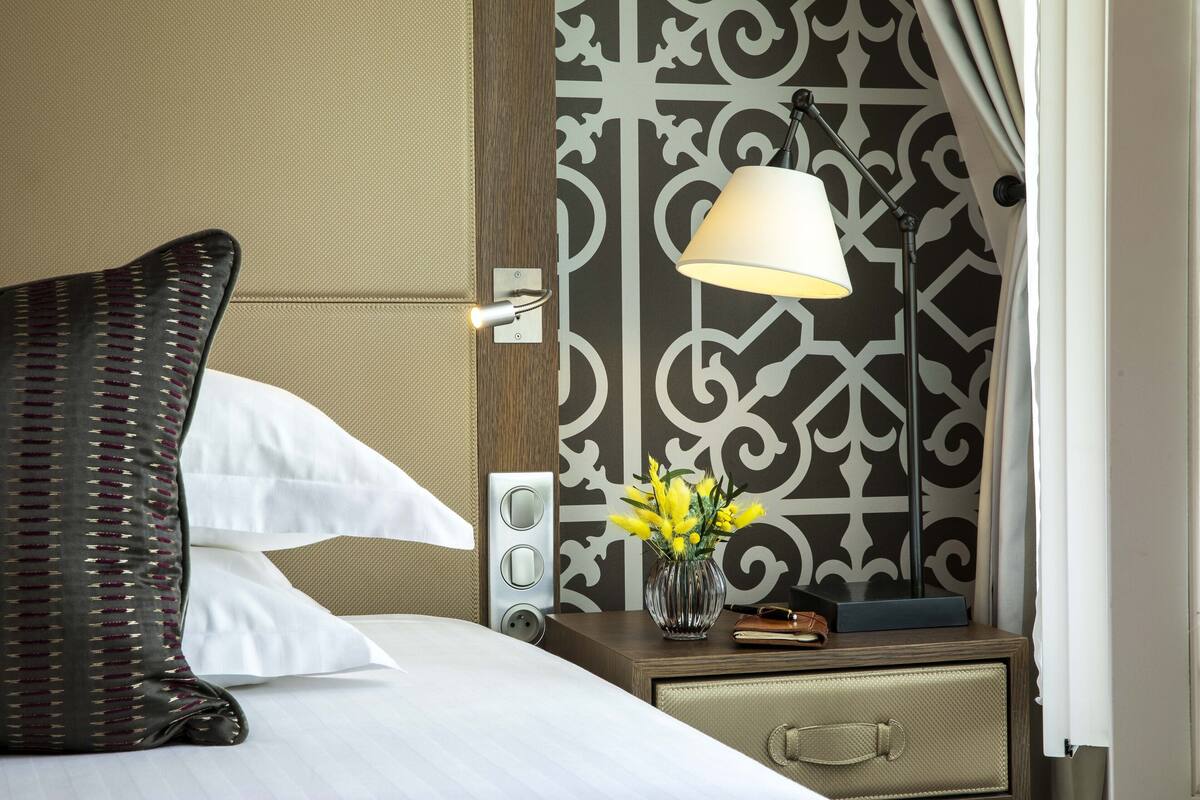 A hotel room with a bed, a nightstand, a lamp, a vase with flowers, and a decorative wall panel.