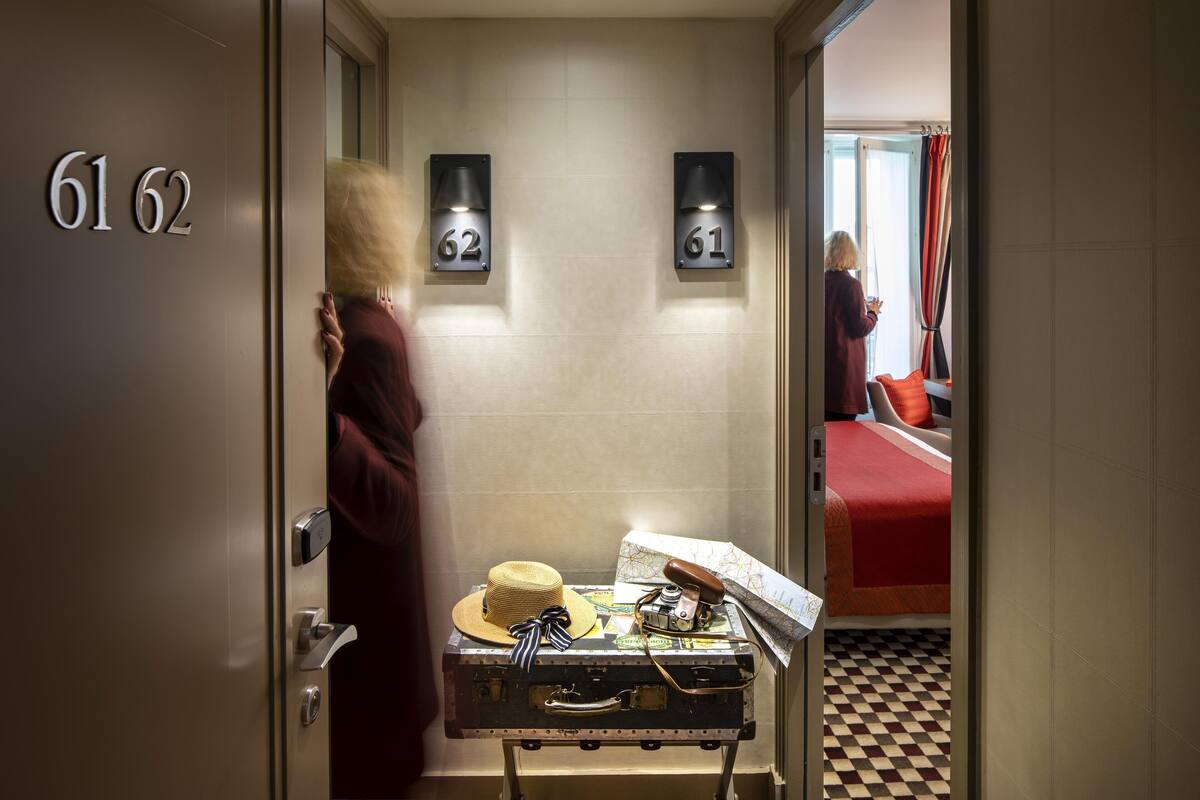 A hotel room with a checkered floor, a suitcase with a hat and camera, and a bed with a red blanket.