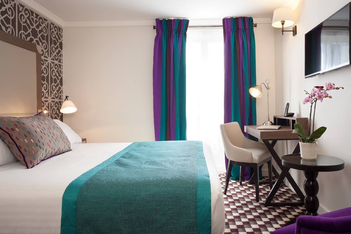 A hotel room with a bed, a desk with a chair, a small table, and a potted plant.