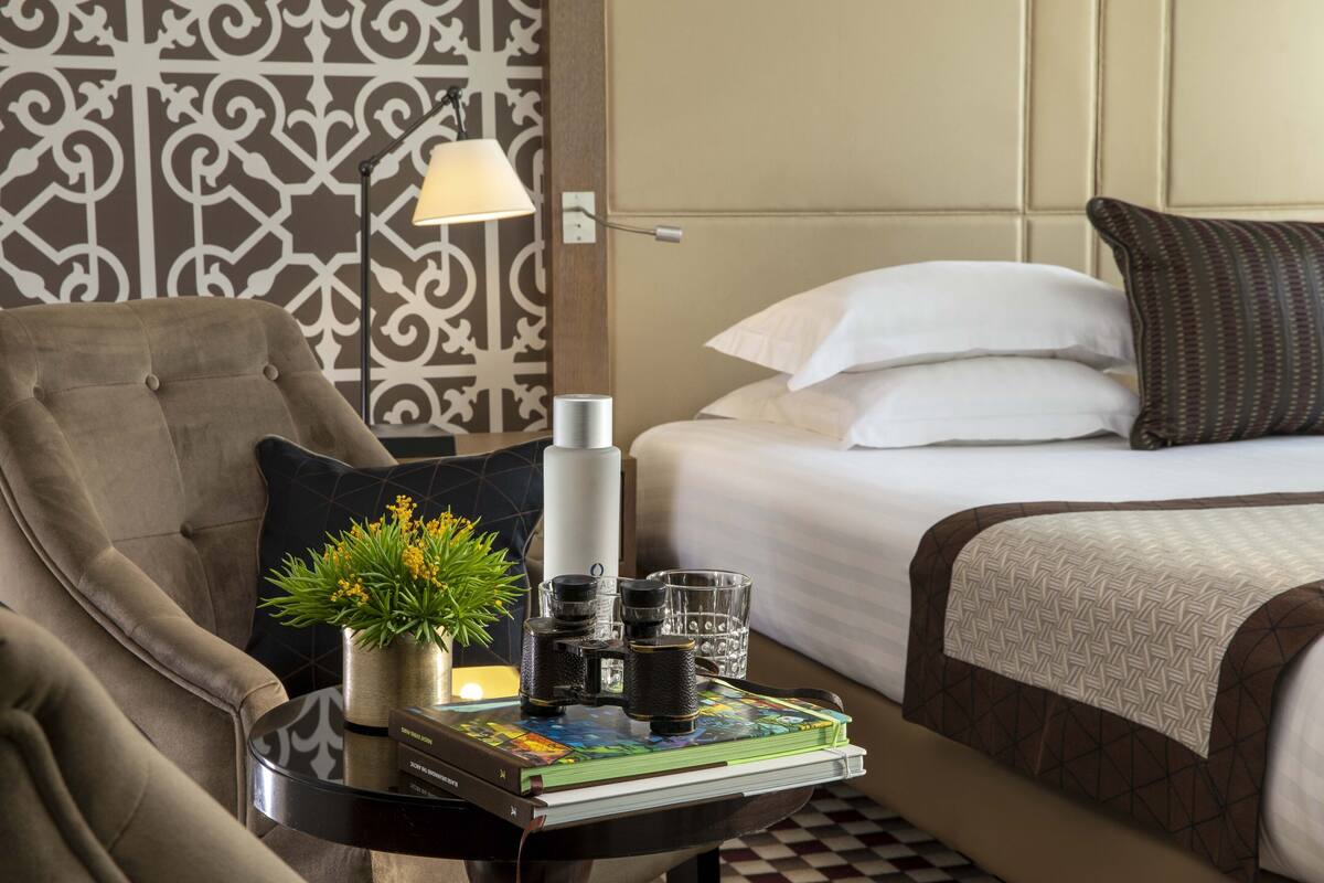 A hotel room with a bed, a chair, a side table with a lamp, a decorative wall, and a small plant.