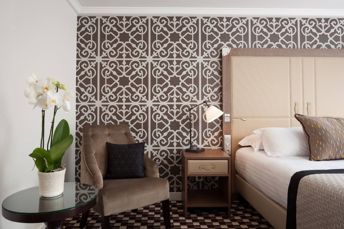 A hotel room with a bed, a chair, a nightstand, a lamp, and a potted plant.