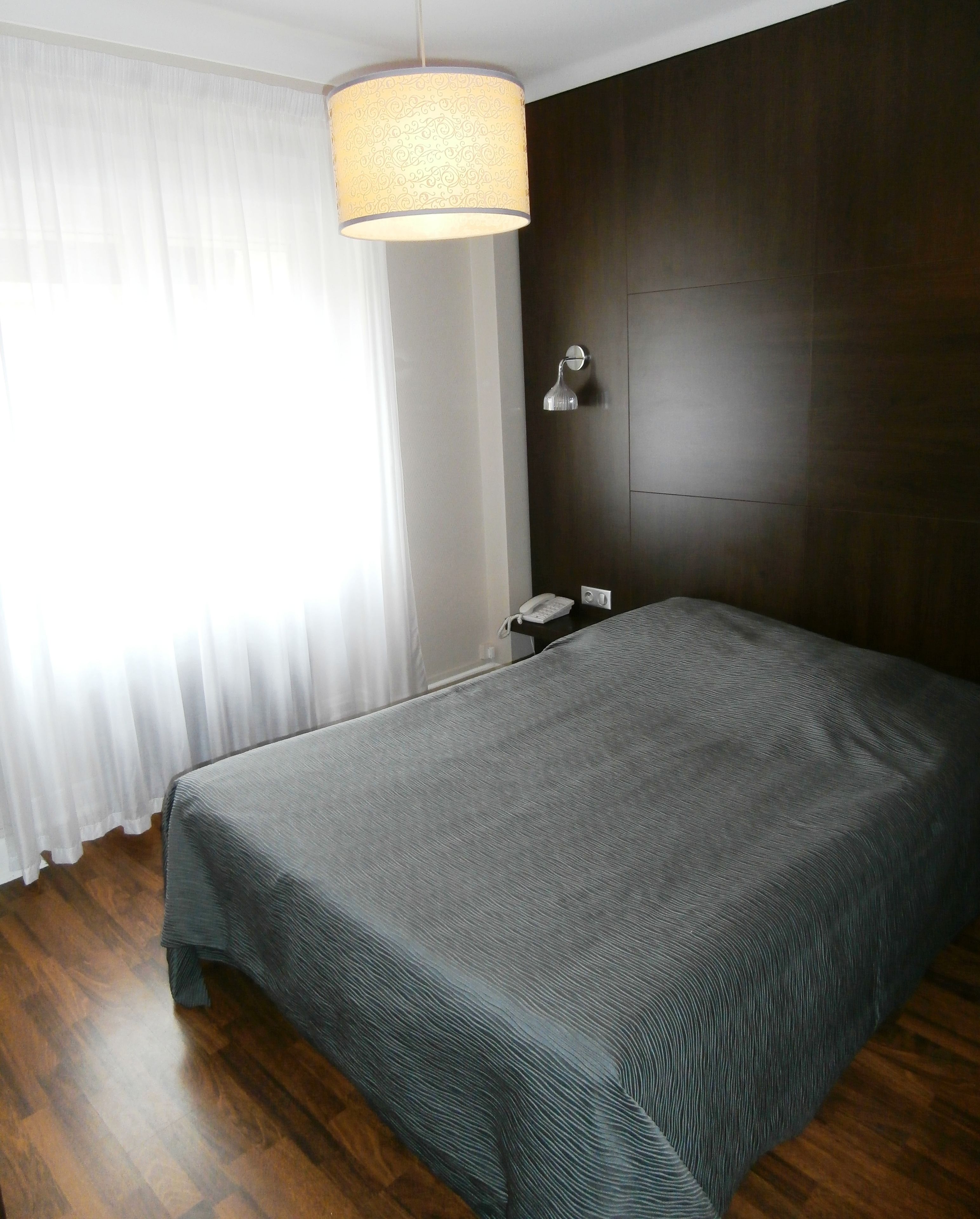 Un dormitorio con una cama grande, una lámpara de noche, un teléfono y una ventana con cortinas translúcidas.