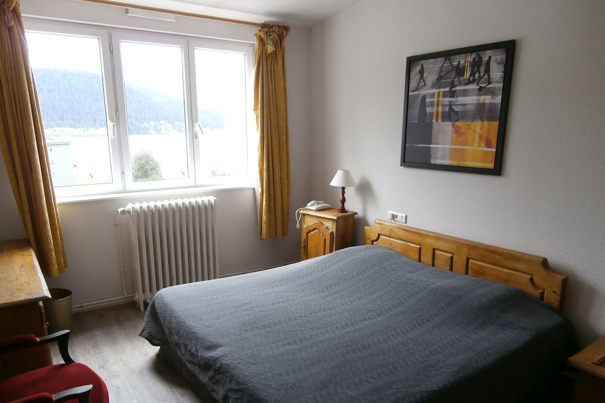 Ett hotellrum med en säng, ett skrivbord med en lampa, en stol, ett element och ett fönster med gardiner.