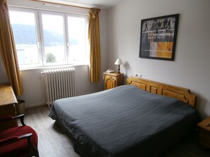 Habitación de hotel con cama, escritorio con lámpara, una silla, radiador y ventana con cortinas.