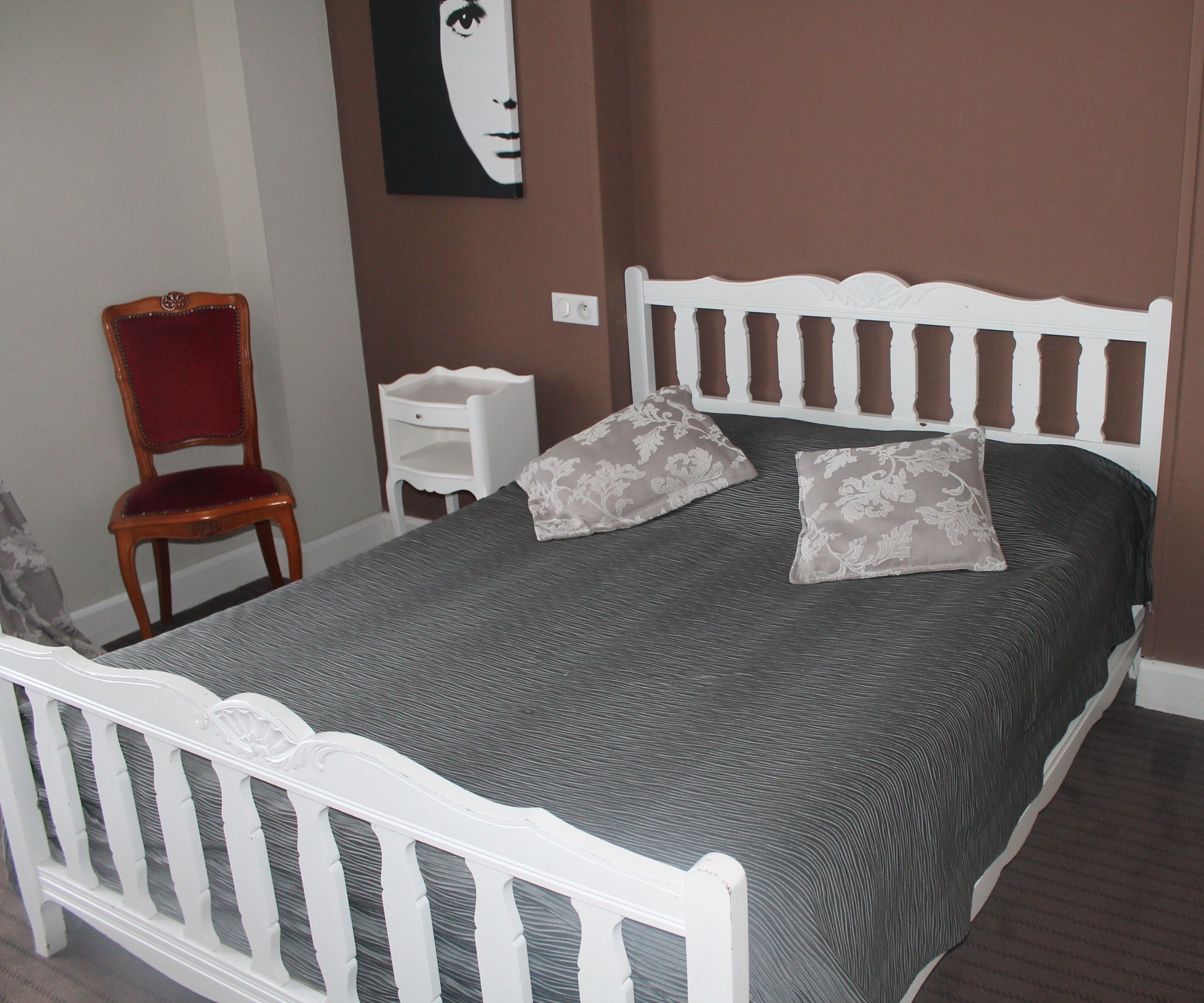 Un dormitorio con estructura de cama blanca, ropa de cama gris, dos almohadas decorativas, una silla de madera y una mesita de noche pequeña.
