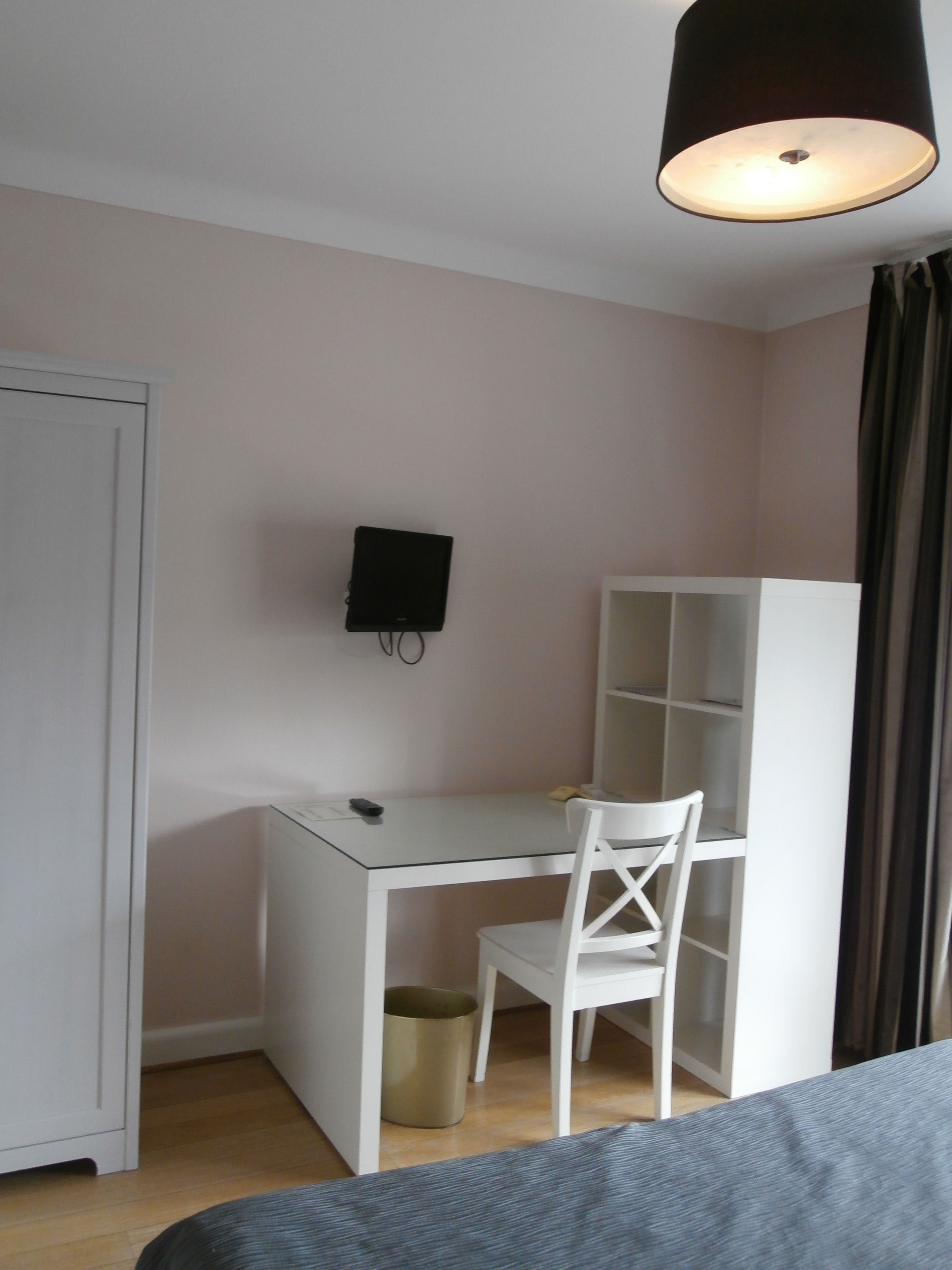 Ett litet, välordnat sovrum med en säng, ett skrivbord med en stol, en tv som är monterad på väggen och en garderob.