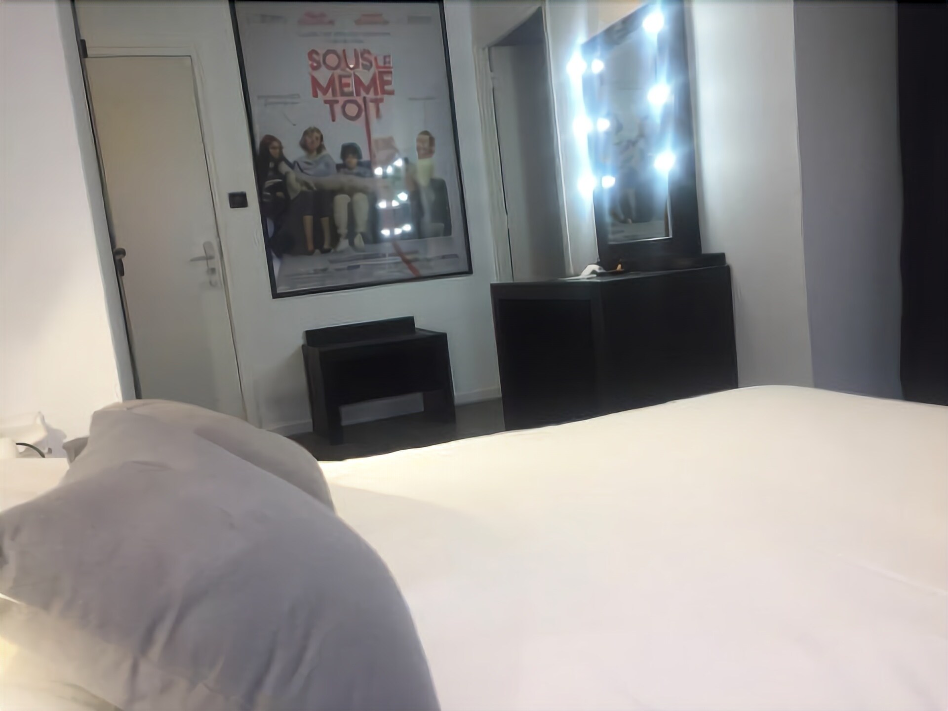 A hotel room with a bed, a small table, a mirror with lights, and a poster on the wall.