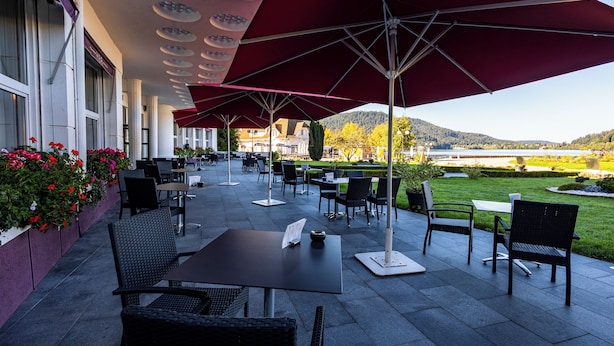 An outdoor patio with tables and chairs, red umbrellas, and a view of a lake and mountains. - Hôtel Beau Rivage (Gerardmer)