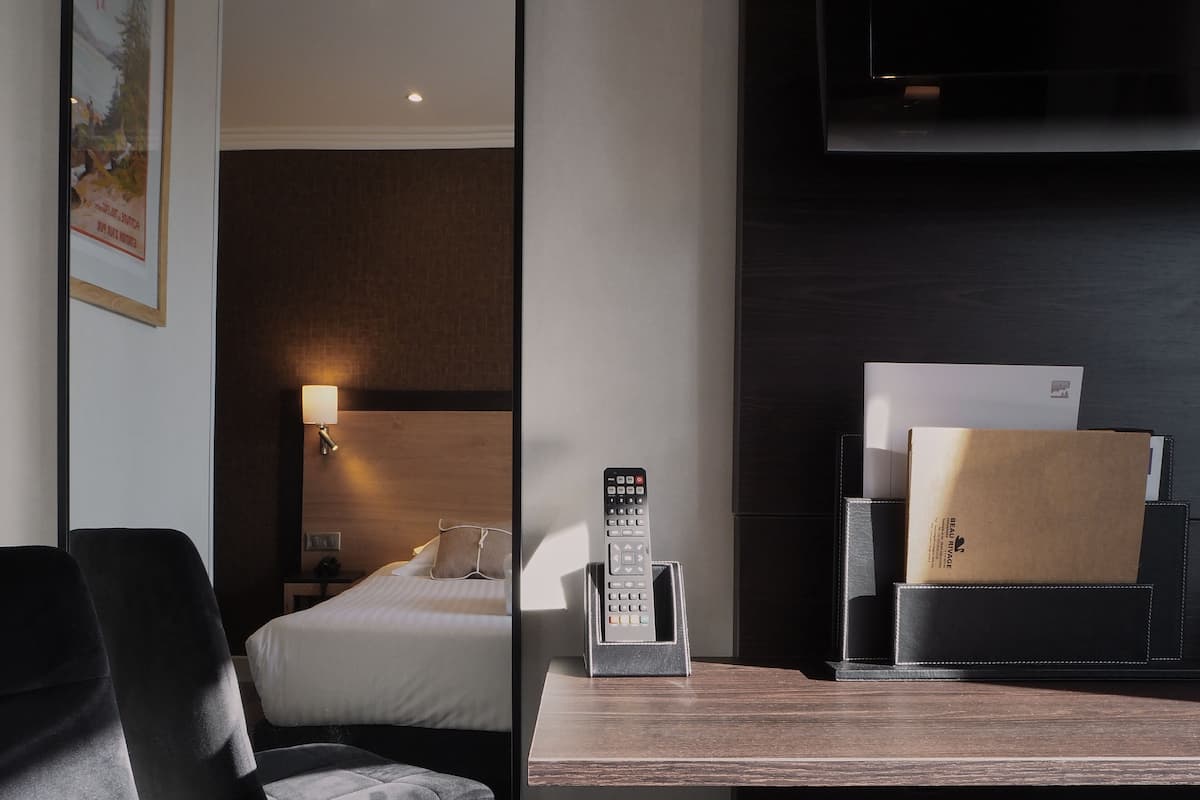 A hotel room with a bed, a desk with a remote control, a chair, and a bedside table with a lamp and a box.