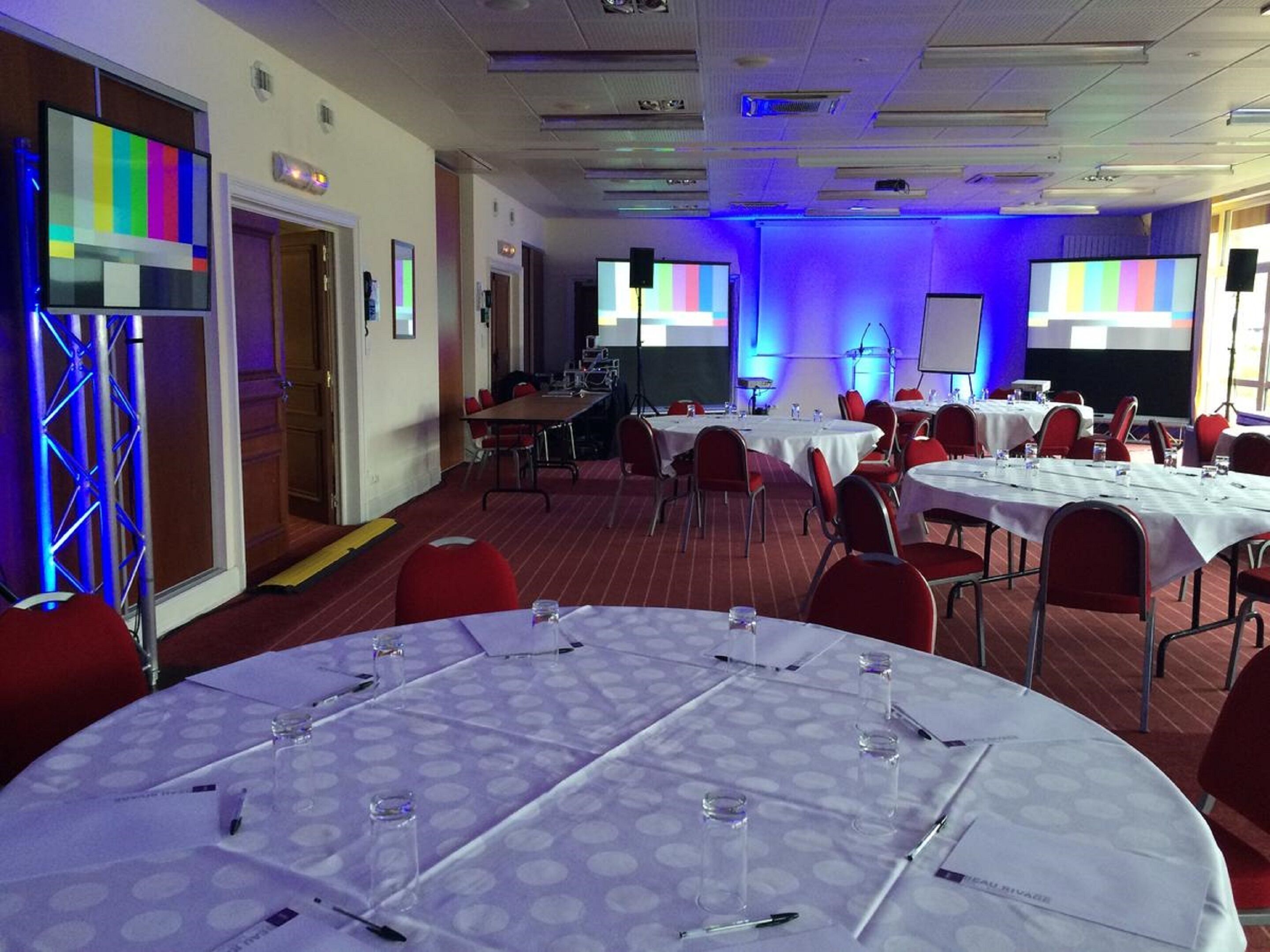 A conference room with round tables, red chairs, and a stage with a projector.