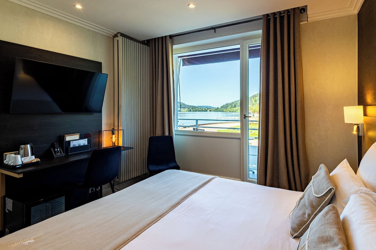 A hotel room with a large bed, a desk with a lamp, a TV, and a view of a lake.