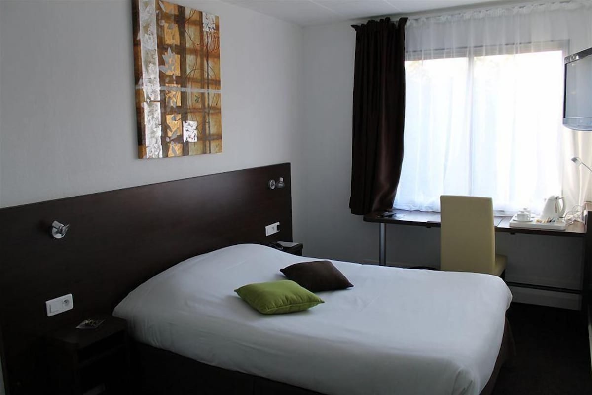 A hotel room with a large bed, a desk with a chair, a window with curtains, and a wall-mounted TV.