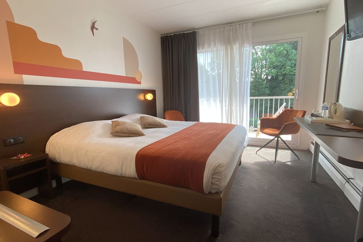 A hotel room with a bed, a desk, a chair, a TV, and a balcony with curtains.
