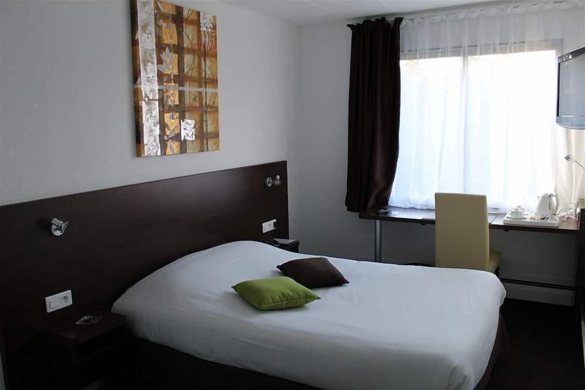 A hotel room with a large bed, a desk with a chair, a window with curtains, and a wall-mounted TV.