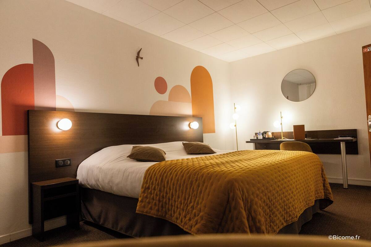 A hotel room with a bed, bedside tables, a desk, a chair, a mirror, and a wall with abstract art.