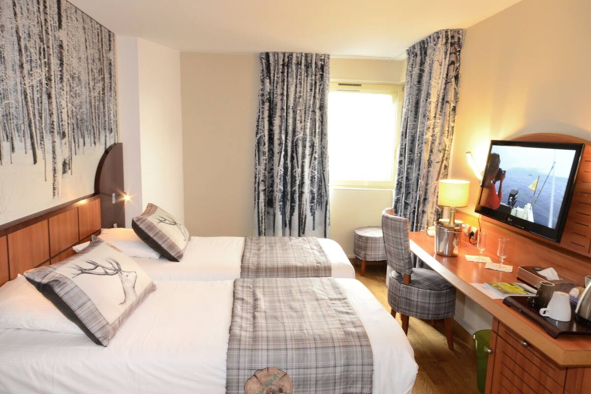 A hotel room with two beds, a desk with a TV, a small seating area, and a large window with curtains.