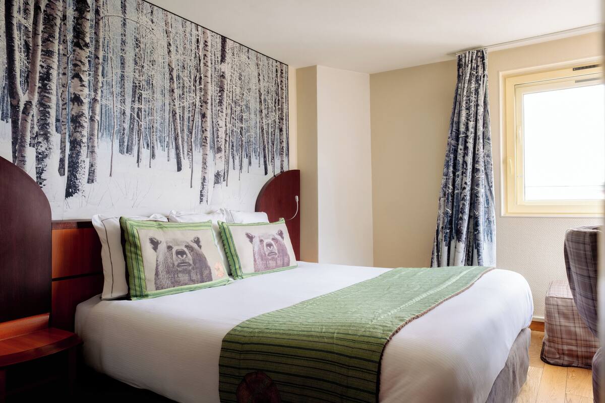 A hotel room with a large bed, a headboard with a bear design, a green striped blanket, and a forest-themed wall mural.