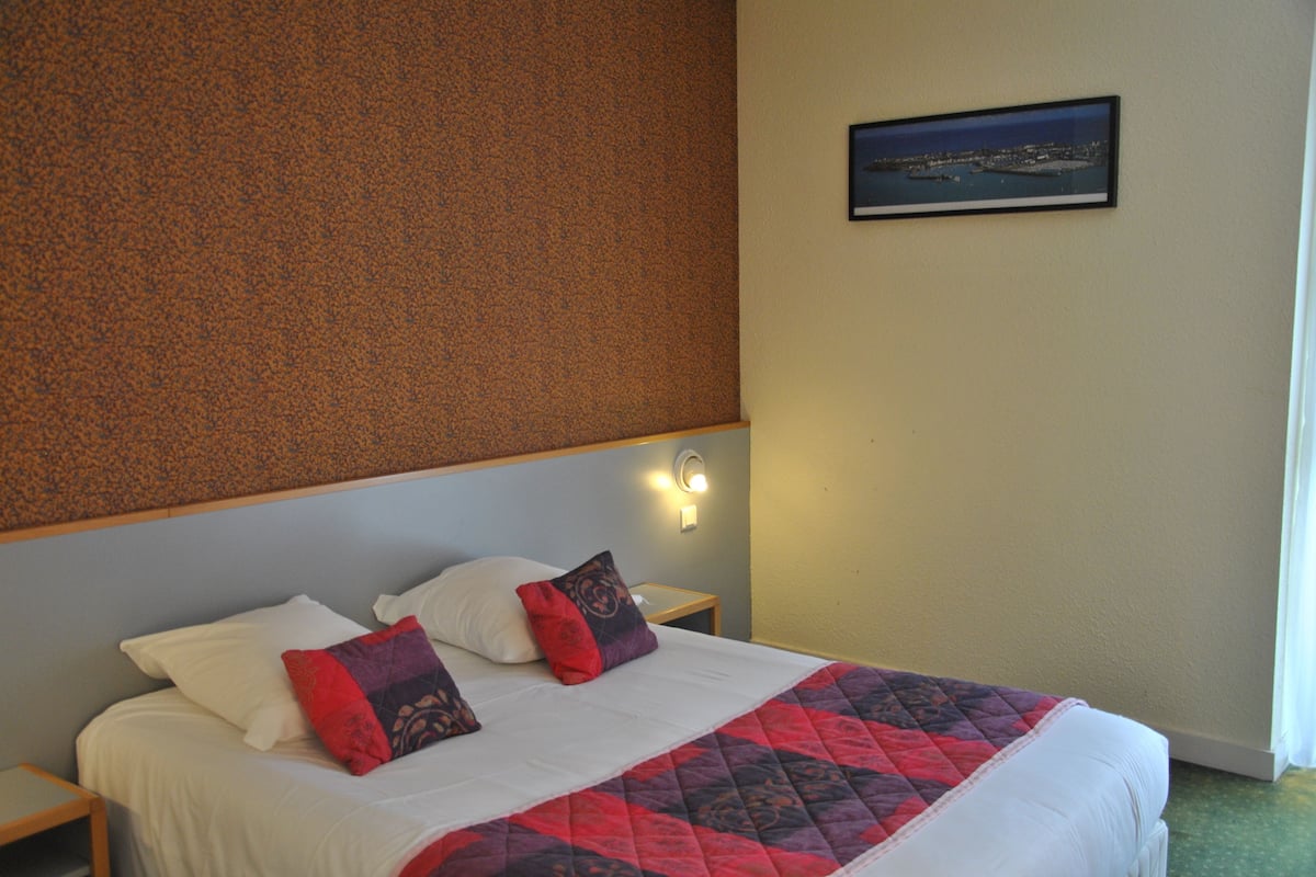 A hotel room with a bed, two pillows, a nightstand, a wall-mounted light, and a framed picture on the wall.