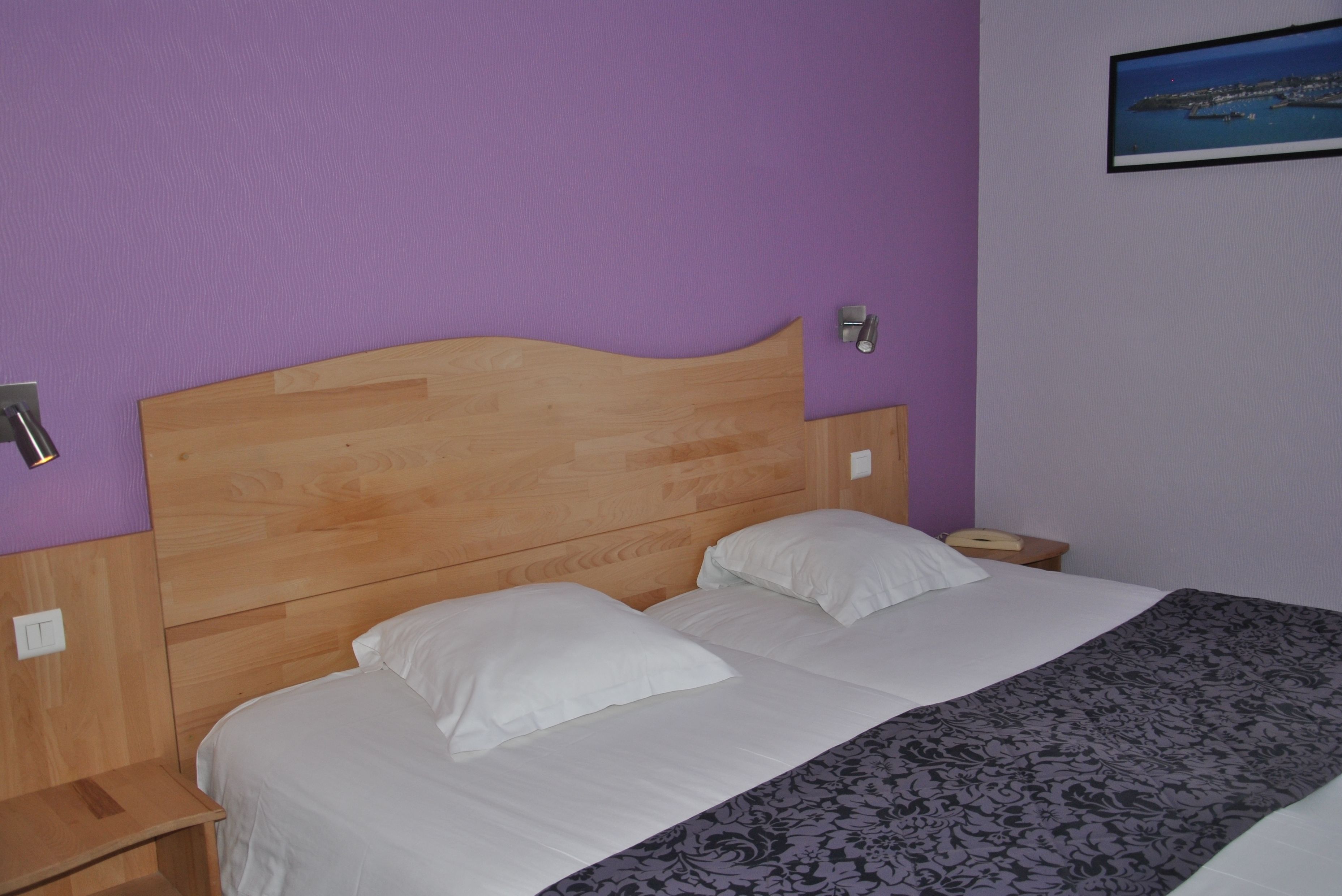 Un dormitorio con cabecera de madera, dos almohadas blancas, colcha estampada y una mesita de noche con lámpara.