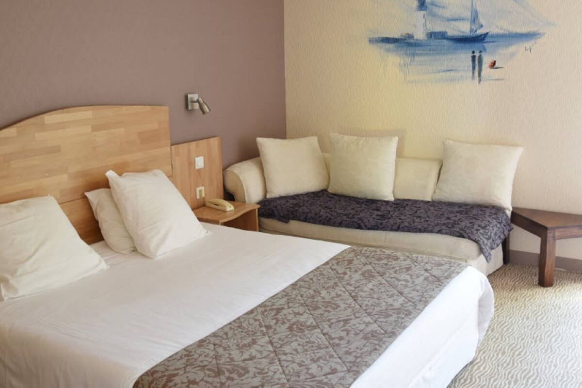 A hotel room with a large bed, a smaller sofa, a bedside table with a lamp, and a painting on the wall.